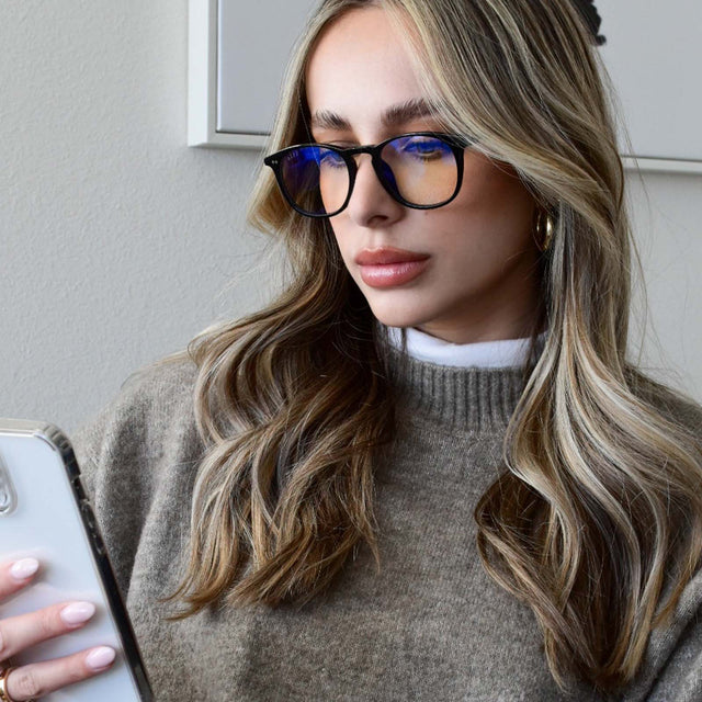lifestyle - blonde female checking her phone wearing diff eyewear jaxson square glasses with a black acetate frame and blue light technology lenses